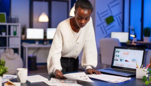 stressed-african-manager-woman-working-with-financial-documents-standing-desk-checking-graphs-holding-papers-reading-raports-late-night-start-up-office-doing-overtime-respect-deadline